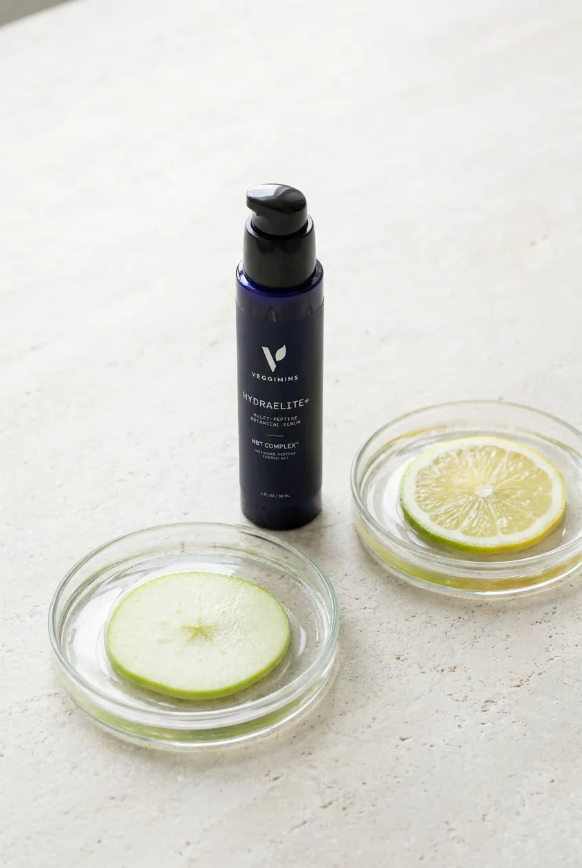 The HydraElite+ bottle beside apple and citrus slices in glass dishes — the two botanical cell cultures
